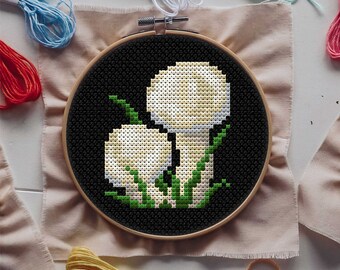 May include: Cross-stitch embroidery hoop with a black background and two white mushrooms with green leaves. The mushrooms are stitched in a simple, pixelated style.