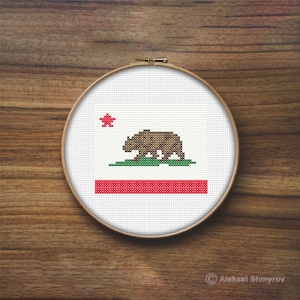 May include: A white embroidery hoop with a cross-stitch design of the California state flag. The flag features a red star, a brown bear walking on green grass, and a red stripe on a white background.
