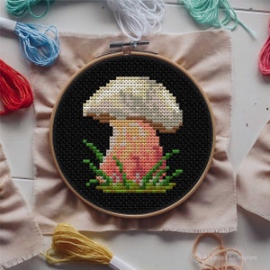 May include: A cross-stitch embroidery hoop with a black background and a colorful mushroom design. The mushroom has a white cap, a pink stem, and green leaves around the base.