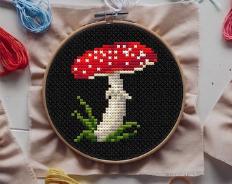 May include: A red and white mushroom with white spots on a black background. The mushroom is embroidered on a piece of fabric in a wooden embroidery hoop.
