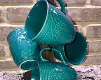 May include: A set of four turquoise ceramic mugs with ornate floral patterns, hanging on a dark wooden mug rack. The mugs have curved handles and are displayed against a brick wall.