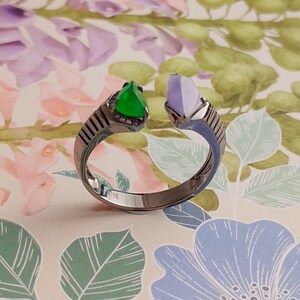Könnte beinhalten: Ein silberner Ring mit einer grünen und violetten Edelsteinbesetzung. Der Ring ist offen und hat ein strukturiertes Band.