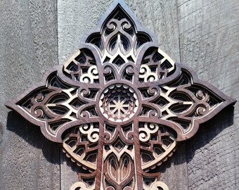 May include: A layered wooden cross with intricate, symmetrical designs. The cross features a central circular element and detailed patterns in shades of brown and tan. The artwork is mounted on a wooden plank background.