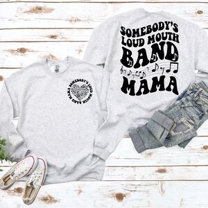 May include: Light gray sweatshirt with the text "Somebody's Loud Mouth Band Mama" in black. The front of the sweatshirt has a heart-shaped design made of musical notes. The back of the sweatshirt has the same text in a larger font with musical notes.
