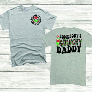 May include: Two gray t-shirts with a green Grinch character wearing a Santa hat. The front shirt has the text "Somebody's Grinch Daddy" in a circle. The back shirt has the text "Somebody's Grinch Daddy" in a larger font.
