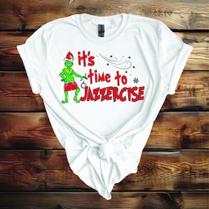 May include: White t-shirt with a green Grinch character wearing a Santa hat and red shorts. The text "It's time to Jazzercise" is printed in red letters.