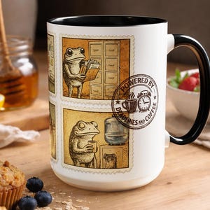 Peut inclure: Mug en céramique blanche avec une anse et un bord noirs. Le mug présente un design fantaisiste de grenouilles dans un style de timbre vintage, avec le texte "POWERED BY DEADLINES AND COFFEE". Le mug est sur une surface en bois.