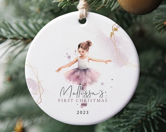 May include: Circular white Christmas bauble with a watercolour illustration of a young ballerina in a pink tutu. The bauble has the text "Melissa's FIRST CHRISTMAS 2023". Gold geometric accents and watercolour splashes complete the design. The bauble hangs from a twine loop.
