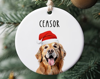 May include: A white ceramic Christmas ornament with a smiling golden retriever wearing a Santa hat. The word "CEASOR" is printed above the dog's head in black text. The ornament is hanging from a natural twine string.