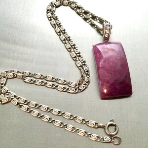 Op de afbeelding: Een zilverkleurige ketting met een rechthoekige, diep magentakleurige stenen hanger. De hanger is bevestigd aan een decoratief zilverkleurig oogje met kleine heldere steentjes. De ketting bestaat uit herhalende S-vormige metalen schakels.