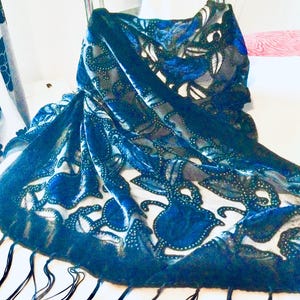 May include: A dark teal scarf with a floral pattern and fringe. The scarf features a detailed design with blue and white accents. The fabric appears to be soft and flowing, with a delicate texture. The scarf is draped, showcasing its shape and design.