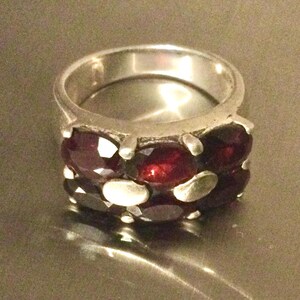 May include: A silver ring featuring two horizontal rows of oval-cut deep red garnet gemstones. The band is constructed from polished silver metal, with the stones held in place by small silver prongs arranged in a repeating pattern around the ring.