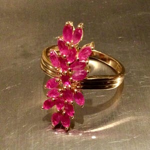 Peut inclure: Une bague dorée ornée d'un groupe de pierres précieuses rubis ovales et vibrantes. Le corps de la bague présente un design multicouche, et les rubis sont disposés en cascade, comme une fleur, créant un bijou luxueux et attrayant.