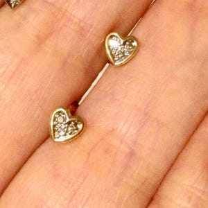 Peut inclure: Une paire de boucles d'oreilles à clous en forme de cœur en or, chacune dotée d'un bord en or poli lisse et d'une section centrale incrustée de petites pierres claires et scintillantes. Les boucles d'oreilles sont petites et ont une silhouette en cœur classique.