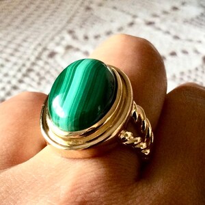 Op de afbeelding: Een gouden ring met een grote, ovaalvormige malachiet edelsteen met opvallende donker- en lichtgroene bandpatronen. De zetting is glad en gepolijst, terwijl de ringband aan de zijkanten een gedraaide, touwachtige textuur vertoont.