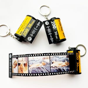 May include: Keyrings featuring film canisters with the text "200 36 exp." and "200 24 EXP." The film canisters are black and yellow. One keyring displays a strip of film with photos of a cat. The keyrings have silver rings.