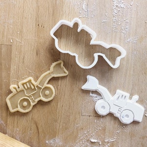 Könnte beinhalten: Zwei weiße Plastik-Ausstecher in Form eines Bulldozers. Ein Ausstecher ist mit hellem braunen Keksteig gefüllt.