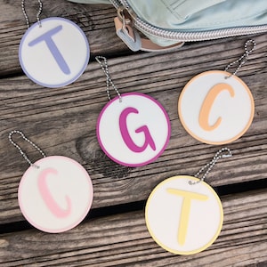 Könnte beinhalten: Fünf runde weiße Anhänger mit bunten Buchstaben, die das Wort "TGCT" buchstabieren. Jeder Anhänger hat eine silberne Kette.