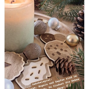 May include: A lit candle next to wooden Christmas ornaments, pine cones, and greenery on a wooden tray. The image includes wooden cookie-shaped decorations and text in German.
