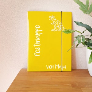 May include: Yellow folder with a white geometric dinosaur design and the text "Postmappe von Maja".