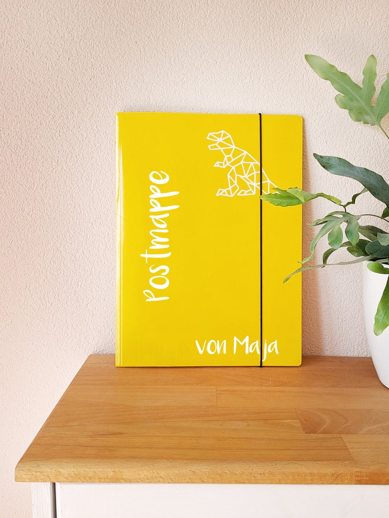 May include: Yellow folder with a white geometric dinosaur design and the text "Postmappe von Maja".