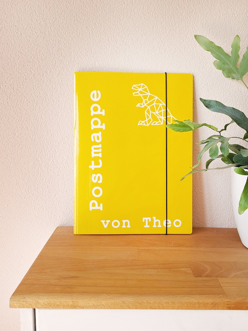 May include: Yellow folder with a white geometric dinosaur design and the text "Postmappe von Theo".