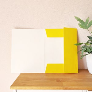 May include: A yellow file folder with a white interior is open on a wooden surface. The folder is sitting on a white cabinet with a wooden knob.
