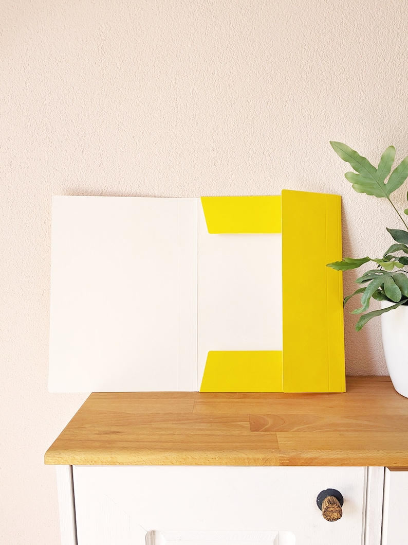 May include: A yellow file folder with a white interior is open on a wooden surface. The folder is sitting on a white cabinet with a wooden knob.
