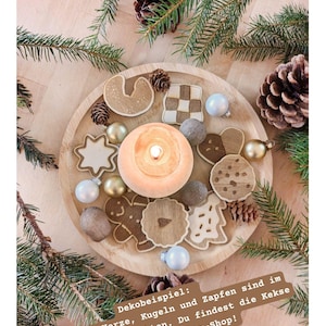 May include: A wooden tray featuring a lit candle, gingerbread biscuits, baubles, and pine cones. The biscuits are in diverse shapes, including stars and gingerbread men. Green pine branches surround the tray, creating a festive holiday display.