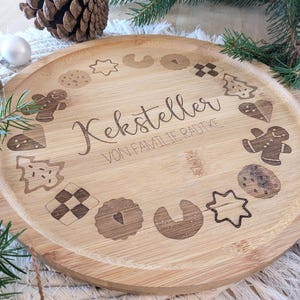 May include: A round, light-brown wooden serving tray with engraved cookie designs and the text "Keksteller VON FAMILIE RADTKE". The tray features various cookie shapes, including gingerbread men and stars, perfect for holiday gatherings.
