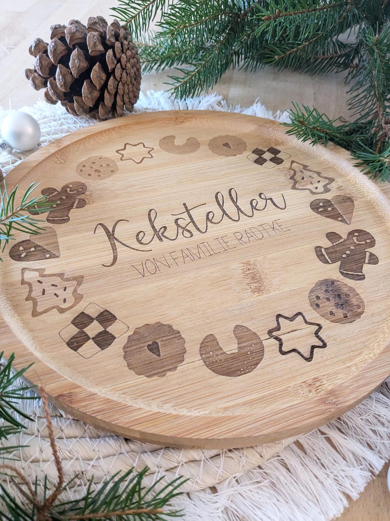 May include: A round, light-brown wooden serving tray with engraved cookie designs and the text "Keksteller VON FAMILIE RADTKE". The tray features various cookie shapes, including gingerbread men and stars, perfect for holiday gatherings.