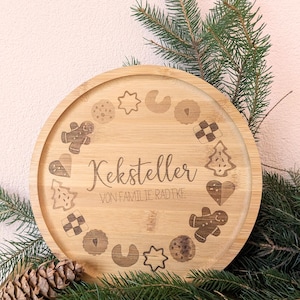 May include: A circular bamboo serving tray with engraved cookie designs and the text "Keksteller VON FAMILIE RADTKE". The tray is surrounded by evergreen branches and a pine cone.