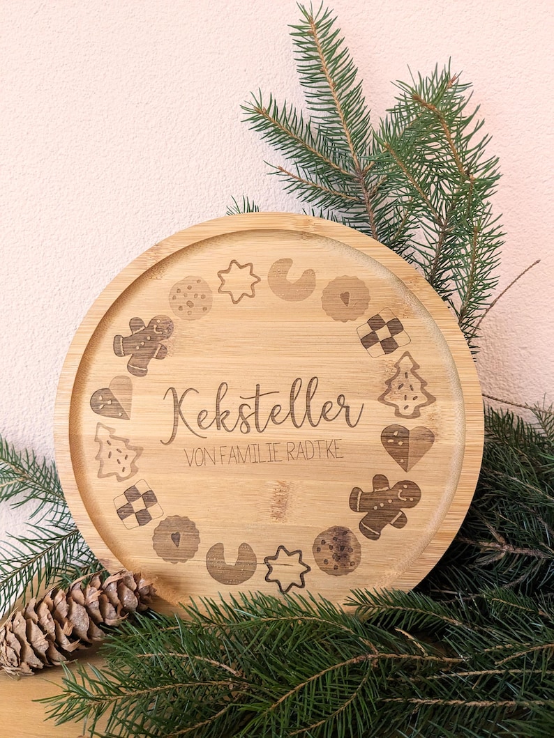 May include: A circular bamboo serving tray with engraved cookie designs and the text "Keksteller VON FAMILIE RADTKE". The tray is surrounded by evergreen branches and a pine cone.