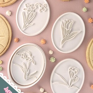 Könnte beinhalten: Ein Set aus vier weißen, ovalen Keksstempeln mit floralen Designs, darunter Narzissen, Tulpen und Schneeglöckchen. Die Stempel sind auf einer rosa Oberfläche mit dekorierten Keksen und kleinen, bunten Süßigkeiten angeordnet.