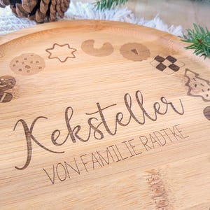 May include: A round, light brown bamboo serving tray with etched cookie designs around the edge. The tray has the words "Keksteller" and "VON FAMILIE RADTKE" etched into the wood. A pine cone and evergreen branches are in the background.