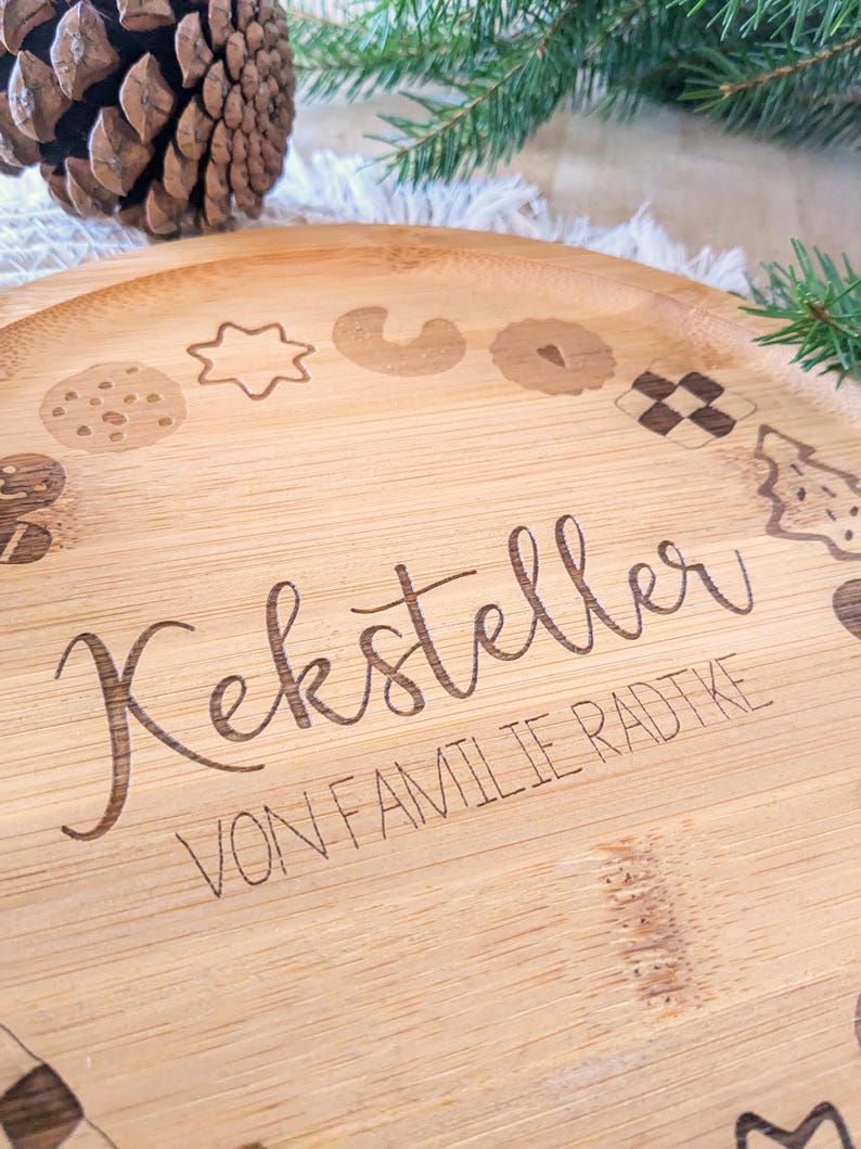 May include: A round, light brown bamboo serving tray with etched cookie designs around the edge. The tray has the words "Keksteller" and "VON FAMILIE RADTKE" etched into the wood. A pine cone and evergreen branches are in the background.