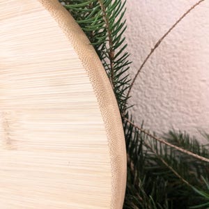 May include: A close-up shot of a light-coloured, round bamboo cutting board with a textured edge. The board is partially framed by green pine needles and a thin, brown twig, suggesting a natural, rustic theme. The board's surface shows the natural grain of the bamboo.