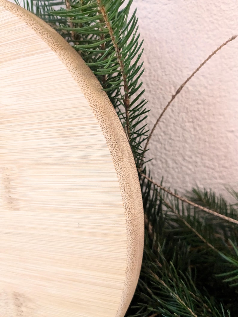 May include: A close-up shot of a light-coloured, round bamboo cutting board with a textured edge. The board is partially framed by green pine needles and a thin, brown twig, suggesting a natural, rustic theme. The board's surface shows the natural grain of the bamboo.