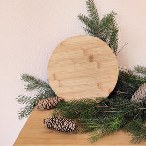 May include: A circular bamboo chopping board, approximately 25 cm in diameter, is surrounded by green pine branches and pine cones. It rests on a wooden surface, with a light pink wall in the background.