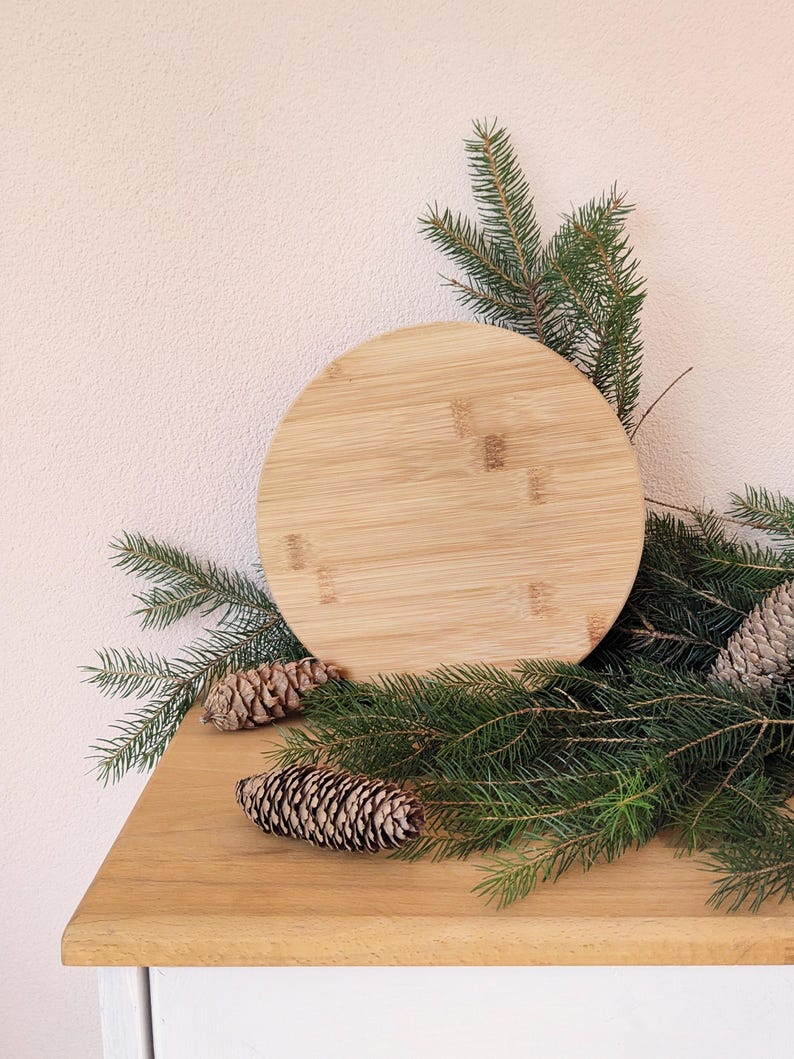 May include: A circular bamboo chopping board, approximately 25 cm in diameter, is surrounded by green pine branches and pine cones. It rests on a wooden surface, with a light pink wall in the background.