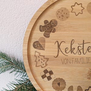 May include: A circular bamboo serving tray with engraved cookie designs, such as a gingerbread man, star, and heart. The tray features the words "Kekste" and "VON FAMILIE". Green pine needles and a pine cone are in the foreground.