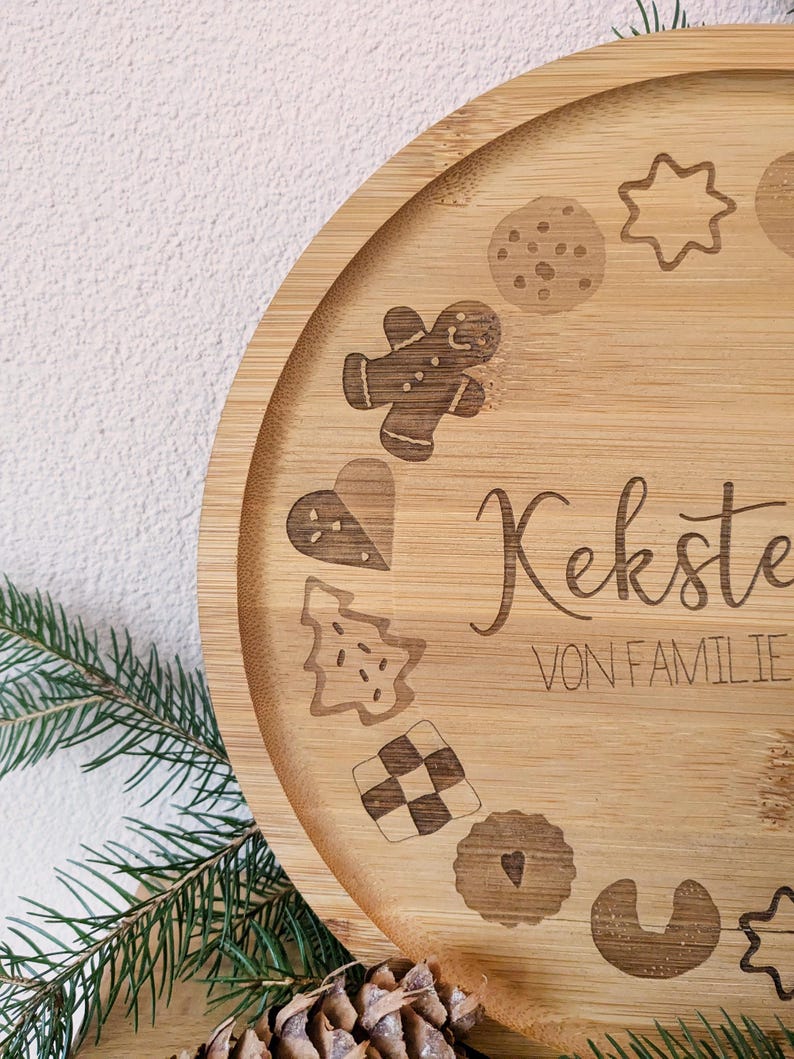 May include: A circular bamboo serving tray with engraved cookie designs, such as a gingerbread man, star, and heart. The tray features the words "Kekste" and "VON FAMILIE". Green pine needles and a pine cone are in the foreground.