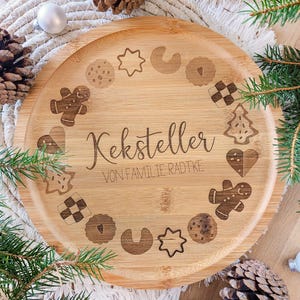 May include: A round, light brown bamboo serving tray with engraved cookie designs and the text "Keksteller VON FAMILIE RADTKE". The tray is surrounded by pine needles, pine cones, and white baubles, suggesting a festive theme.