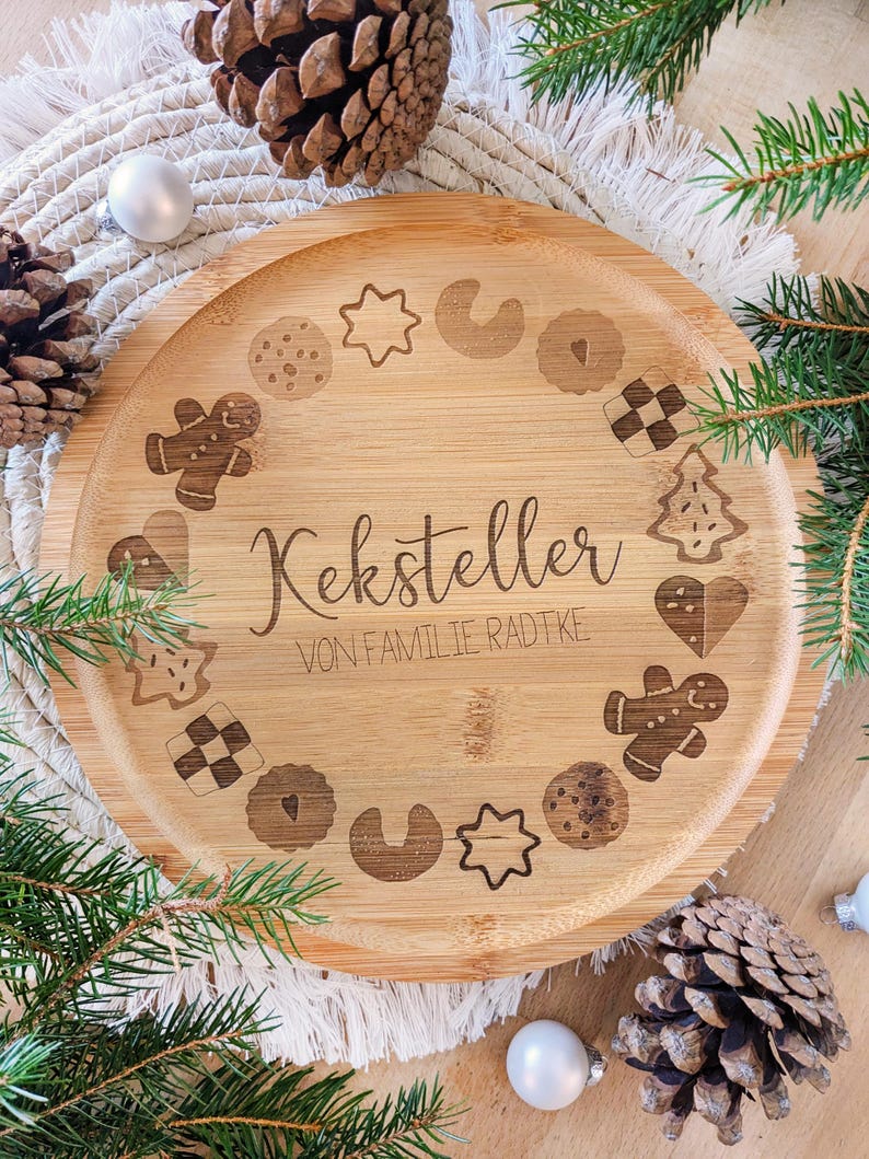 May include: A round, light brown bamboo serving tray with engraved cookie designs and the text "Keksteller VON FAMILIE RADTKE". The tray is surrounded by pine needles, pine cones, and white baubles, suggesting a festive theme.