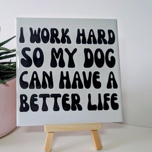 May include: A white square tile with the text "I WORK HARD SO MY DOG CAN HAVE A BETTER LIFE" in black, displayed on a small wooden easel. The tile is in front of a white background, with a plant in a pink pot visible on the left.