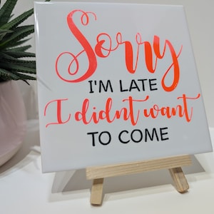 May include: A white ceramic tile with the text "Sorry I'm late I didn't want to come" in black and orange lettering. The tile is on a small wooden easel.