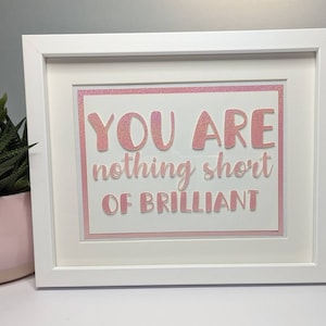 Puede incluir: Una impresión enmarcada con las palabras "YOU ARE nothing short OF BRILLIANT" en letras rosas con purpurina. El marco blanco rodea un passe-partout blanco y un borde rosa con purpurina. Una planta suculenta en una maceta rosa es visible en el fondo.