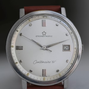 May include: A vintage Eterna-Matic Centenaire '61 wristwatch with a silver case and a white dial. The dial features silver hands, hour markers, and the date at the 3 o'clock position. The watch has a brown leather strap.