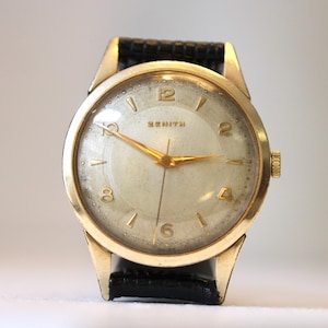 May include: A vintage Zenith wristwatch with a gold-tone case and a cream-coloured face. The watch features gold hands and hour markers, a black leather strap, and the word "ZENITH" printed on the face.