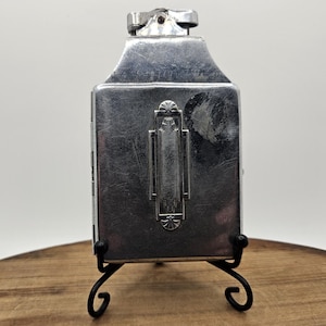 May include: A vintage silver-toned lighter with a rectangular body and a decorative front panel. The lighter is displayed on a black metal stand with curved legs, set against a wooden surface.